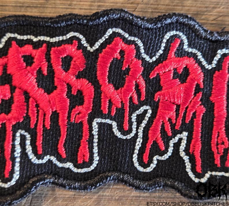 Puede incluir: Un parche bordado rectangular con fondo negro y letras rojas estilizadas que parecen gotear. El texto est&aacute; delineado con un hilo blanco fino, y todo el parche tiene un borde grueso cosido en negro con un contorno ondulado.