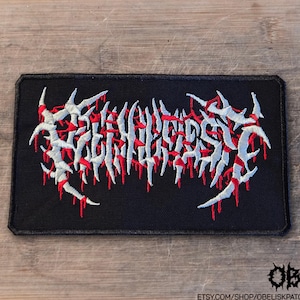 May include: A rectangular black embroidered patch featuring white, jagged, thorn-like lettering with red accents that resemble dripping blood. The design is centered on the black fabric background, which has a stitched border around the edges.
