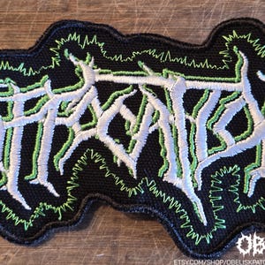 May include: A black embroidered patch featuring white, stylized, jagged lettering outlined in bright green thread. The design has a dense, textured appearance with a black border and a jagged green stitch pattern surrounding the central text.
