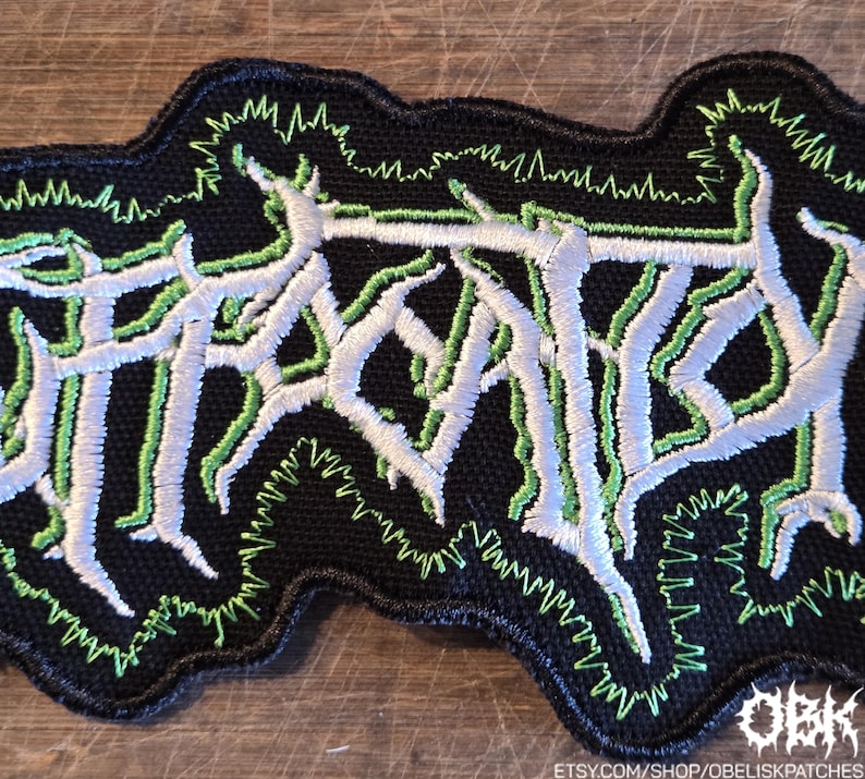 May include: A black embroidered patch featuring white, stylized, jagged lettering outlined in bright green thread. The design has a dense, textured appearance with a black border and a jagged green stitch pattern surrounding the central text.