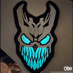 以下が含まれることがあります： 光る青い目と歯を持つ、様式化されたスカルデザインの刺繍パッチ。パッチは黒い縁取りと薄い灰色のスカルです。「OBK」の文字とEtsyショップのURLが表示されています。