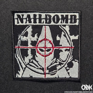 Pode incluir: Patch quadrado preto com a palavra "NAILBOMB" em branco. O design apresenta uma mira vermelha e cinza com uma figura estilizada. O patch é bordado com fio branco e cinza.