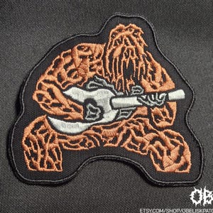 Puede incluir: Un parche bordado negro que presenta una criatura humanoide peluda de color cobre tocando una guitarra eléctrica blanca. El diseño está delineado con hilo negro y el parche tiene una forma irregular con una apariencia texturizada y cosida.