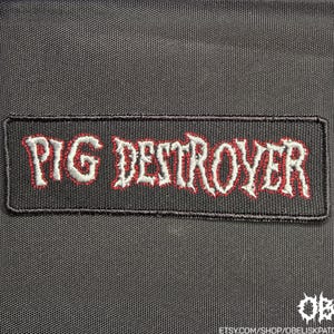 以下が含まれることがあります： 黒い長方形の刺繍ワッペン。細い赤い縁取りが施された白いスタイライズドフォントで「PIG DESTROYER」という文字が刺繍されています。ワッペンには黒い縁があり、織り目の見える黒い布地の上に配置されています。