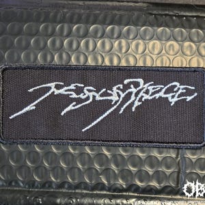 May include: A rectangular black embroidered patch featuring the white text Jesus Piece in a stylized, jagged, and cursive script. The patch has a thick black stitched border and a textured fabric surface, presenting a bold and high-contrast design.