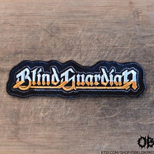 May include: A black embroidered patch featuring the text Blind Guardian in a stylized font. The lettering has a gradient effect, transitioning from a light blue color at the top to a vibrant orange color at the bottom, with a black border.