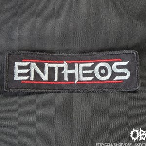 Puede incluir: Un parche rectangular bordado en negro que presenta la palabra ENTHEOS en letras mayúsculas de color gris plateado. El texto está bordeado por finas líneas horizontales rojas encima y debajo de las letras, sobre un fondo de tela negra.