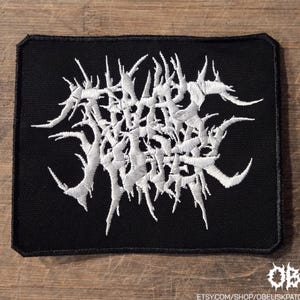 May include: A rectangular black fabric patch featuring white embroidered text in a stylized, jagged, and illegible font. The patch has a stitched border and includes the small white text "OBK" and "etsy.com/shop/obeliskpatches" in the corner.