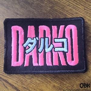 May include: Embroidered patch with the word "DARKO" in bright pink, with Japanese characters in light blue. The patch is rectangular with a black background and a stitched border. The patch is likely for clothing or accessories.