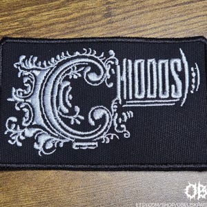 May include: Black rectangular patch with white embroidered text and ornate design. The word "CHIODOS)" is displayed in a stylized font. The letter "C" is large and decorative. The patch is on a wooden surface.