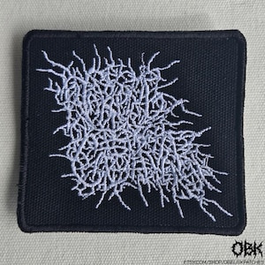 Könnte beinhalten: Ein quadratischer, schwarzer Stickaufnäher mit einem dichten, komplizierten Design aus hellgrauem Garn. Die zentrale Grafik besteht aus komplexen, gezackten und miteinander verbundenen Linien, die ein stilisiertes, unleserliches Textmuster bilden.