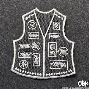 May include: A black embroidered patch shaped like a vest, featuring white stitched borders and various white graphic designs including circular and rectangular motifs. The design includes intricate patterns and symbols across the front surface.