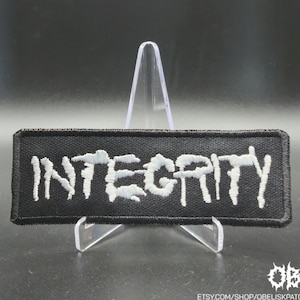 May include: Black rectangular patch embroidered with the word "INTEGRITY" in white, stylized lettering. The patch has a black border and is displayed on a clear stand. The Etsy shop name "OBK" is in the bottom right corner.