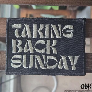 May include: A rectangular black embroidered patch featuring the text TAKING BACK SUNDAY in light gray, stylized, block-style lettering. The patch has a stitched border and a textured fabric surface, with the letters clearly visible against the dark background.