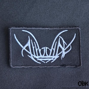 Puede incluir: Un parche rectangular de tela negra con texto bordado en blanco en una caligrafía estilizada y angular. El bordado está centrado en el parche, que tiene un borde cosido. El diseño consiste en líneas nítidas y fluidas que forman letras.