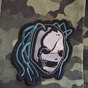 Puede incluir: Parche bordado con un diseño de rostro estilizado con máscara, en tonos grises y negros, con detalles turquesa. El parche se encuentra sobre un fondo de camuflaje, creando un contraste llamativo. Ideal para ropa o accesorios.