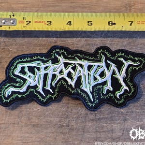 Pode incluir: Um emblema bordado preto com o texto branco Suffocation numa fonte estilizada e irregular com um contorno verde brilhante. Uma fita métrica amarela Stanley está posicionada acima do emblema, mostrando um comprimento de cerca de 19 cm.
