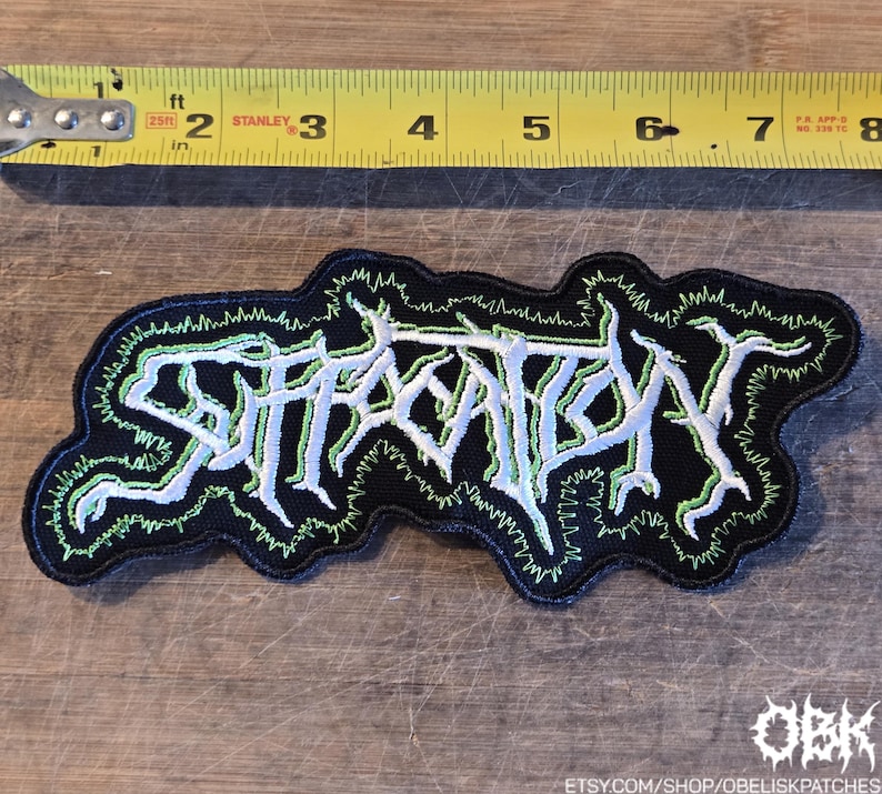May include: A black embroidered patch featuring the white text Suffocation in a jagged, stylized font with a bright green outline. A yellow Stanley measuring tape is positioned above the patch, showing it is approximately 7.5 inches in length.