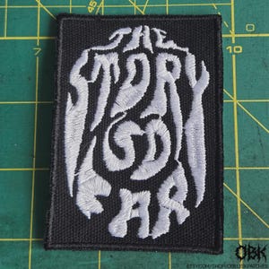 May include: Black rectangular patch with white embroidered text that reads "THE STORY GOD WAR" in a stylized font. The patch has a black border and is likely an iron-on or sew-on patch for clothing or accessories.