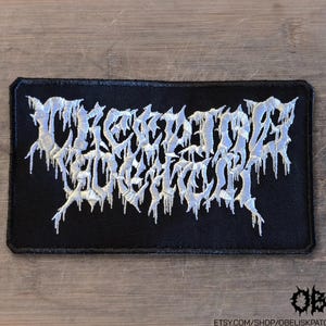 May include: A rectangular black embroidered patch featuring the white text "CREEPING DEATH" in a stylized, dripping, jagged font. The embroidery has a textured appearance with sharp, pointed edges, and the patch has a clean, stitched border.