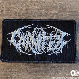 May include: A rectangular black embroidered patch featuring white thread forming a complex, stylized, and illegible text design in the center. The patch has a solid black border and a textured fabric surface, with the text appearing as a dense, intricate, and thorny graphic pattern.