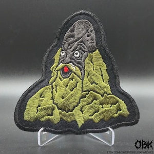 Könnte beinhalten: Ein gestickter Aufnäher mit einer dunkelgrauen, texturierten Kapuzenfigur mit langem, fließendem olivgrünem Bart und Gewand. Die Figur hat kleine weiße Augen und ein leuchtend rotes kreisförmiges Detail am Mund, alles schwarz umrandet.