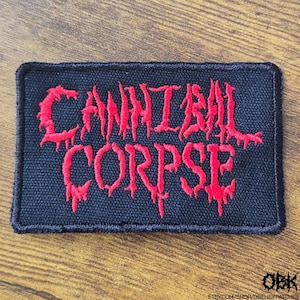 Peut inclure: Un écusson rectangulaire noir avec les mots "CANNIBAL CORPSE" brodés en rouge, dans une police gothique. L'écusson a une bordure noire et est probablement en tissu. Un petit logo et une adresse de site web figurent dans le coin inférieur droit.