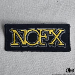 Puede incluir: Parche rectangular negro con la palabra "NOFX" bordada en hilo amarillo y gris. El parche tiene un borde negro y está diseñado para ser cosido o planchado en ropa o accesorios. El parche está sobre una tela gris claro.