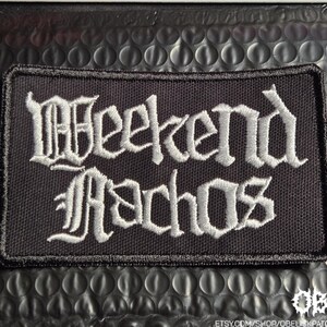 May include: A rectangular black embroidered patch with a thick black stitched border. The patch features the text "Weekend Nachos" in a white, stylized gothic font. The embroidery is dense and covers the entire surface of the fabric base.