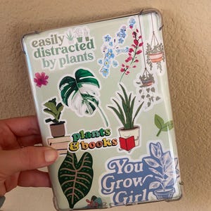 Puede incluir: Una funda de plástico transparente para un lector electrónico con varias pegatinas. Las pegatinas muestran plantas, flores y texto que dice "easily distracted by plants", "plants & books" y "You Grow Girl".