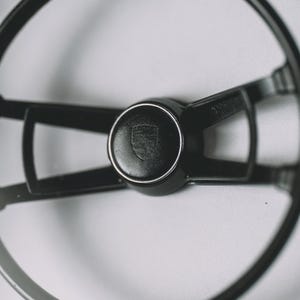 Puede incluir: Primer plano de un volante negro con un emblema central. El volante tiene un borde circular y radios. El emblema es un escudo. La imagen está sobre un fondo blanco, destacando el diseño del volante.