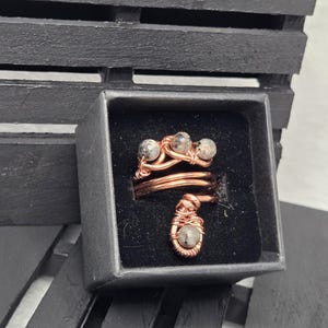 May include: A copper-colored wire-wrapped ring with three small, speckled stones. The ring is in a black box, set against a dark background. The ring's design features a spiral and a dangling stone.
