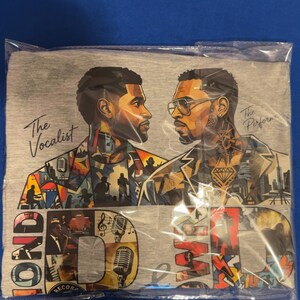 May include: A heather gray t-shirt featuring a graphic print of two men facing each other. The left figure is labeled The Vocalist, and the right is labeled The Performer. The design includes colorful collage elements, microphones, and large block letters.