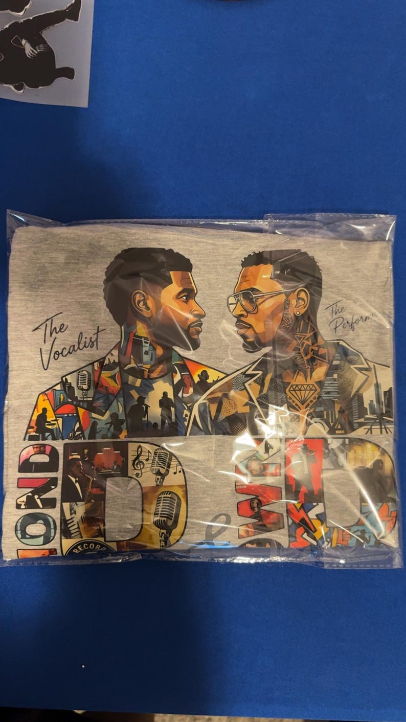 May include: A heather gray t-shirt featuring a graphic print of two men facing each other. The left figure is labeled The Vocalist, and the right is labeled The Performer. The design includes colorful collage elements, microphones, and large block letters.