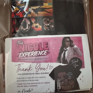 May include: A black t-shirt with a colorful graphic print featuring musical artists, packaged in a clear plastic sleeve with a pink and white thank you card. The card displays the text Tha NICOLE EXPERIENCE and includes a photo of a person.