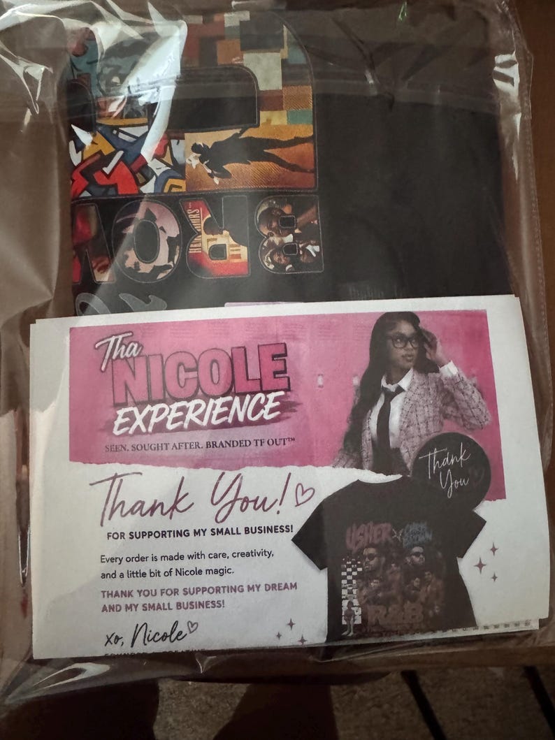 May include: A black t-shirt with a colorful graphic print featuring musical artists, packaged in a clear plastic sleeve with a pink and white thank you card. The card displays the text Tha NICOLE EXPERIENCE and includes a photo of a person.