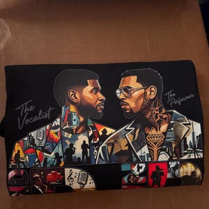 May include: A black t-shirt featuring a graphic illustration of two men facing each other. The left figure is labeled The Vocalist, and the right figure is labeled The Performer. The design includes colorful collage-style imagery of microphones, cityscapes, and musical silhouettes.