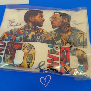May include: A white t-shirt in a clear plastic bag featuring a graphic print of two men facing each other. The text reads The Vocalist and The Performer. The design includes colorful collage elements, musical instruments, microphones, and large letters.