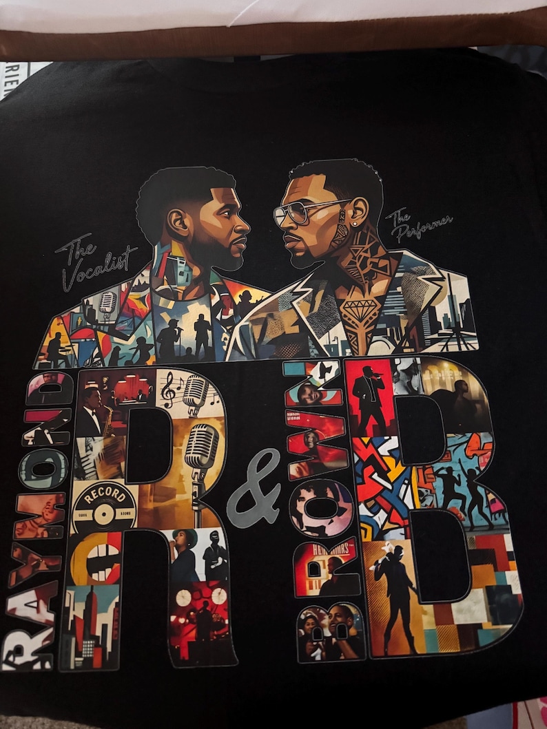 May include: A black t-shirt featuring a graphic print of two men facing each other, labeled The Vocalist and The Performer. Large letters spell out Raymond & Brown, filled with a collage of musical scenes, microphones, and city silhouettes.