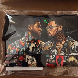 May include: A black t-shirt featuring a graphic illustration of two men facing each other. The left side shows a man labeled The Vocalist with a microphone, while the right side shows a man labeled The Performer wearing glasses. The design includes colorful collage elements of musical scenes and cityscapes.