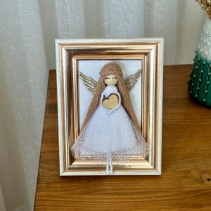 May include: A framed white yarn angel figurine holding a wooden heart. The angel has brown yarn hair and wings. The frame is white with gold trim.