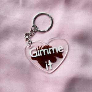 May include: Clear heart-shaped keychain with the words "Gimme it" in white lettering and a brown graphic. The keychain has a silver metal ring and chain.