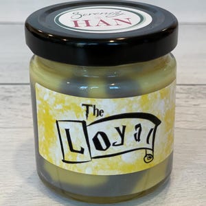 May include: A glass jar with a black lid and a yellow label that says "The Loyal" with a black and white design. The jar is filled with a yellow and gray candle.