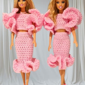 Könnte beinhalten: Zwei Barbie-Puppen in passenden rosa Häkelkleidern. Die Kleider haben ein Crop-Top mit großen Rüschenärmeln und einen Bleistiftrock mit Rüschensaum. Der Text "Evirosa Crochet Dress" steht oben.