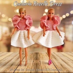 Könnte beinhalten: Zwei Barbie-Puppen in handgefertigten Häkelkleidern. Die Kleider haben ein rosa Oberteil mit Rüschenärmeln und einen weißen, ausgestellten Rock. Der Text "Caelaria Roselle Dress" steht oben. Die Puppen stehen auf einer Holzoberfläche.