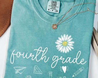 Puede incluir: Una camiseta Comfort Colors turquesa con las palabras "fourth grade" en escritura blanca, un gráfico de margarita e iconos escolares. También se ven un collar dorado y un sombrero marrón.