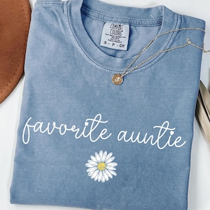 Peut inclure: Un t-shirt bleu avec l'inscription "favorite auntie" en écriture blanche et un motif de marguerite. Un collier en or avec un pendentif est drapé sur le t-shirt. Le t-shirt est de la marque Comfort Colors.