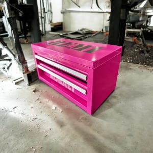 May include: A vibrant pink tool chest with two drawers and a top section. The word "KOBALT" is printed in white on the front of the drawers, and "REMIX" is printed in black on the top. The drawer handles are silver.