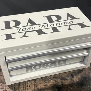 May include: A white metal tool chest with two drawers and silver handles. The top surface features the black text "PAPA" with the name "Jose Moreno" centred horizontally. The word "KOBALT" is printed in grey on the bottom drawer front.