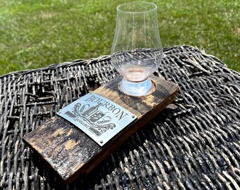 May include: A clear, tulip-shaped bourbon glass rests on a wooden coaster. The coaster features a metal plate with the word "BOURBON" and a decorative design. The wood has a dark, weathered appearance.